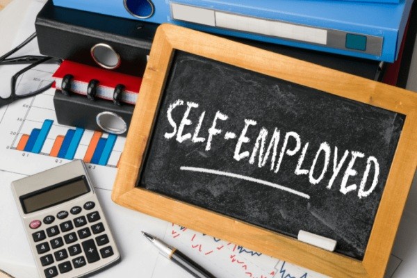 Chalkboard that says _Self-employed_ on table with binders, documents, and calculator