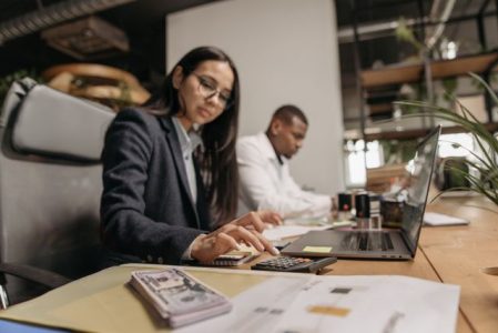 Woman using a calculator alongside a laptop and cash while reviewing finances, illustrating how does an HSA work for tax savings and budgeting